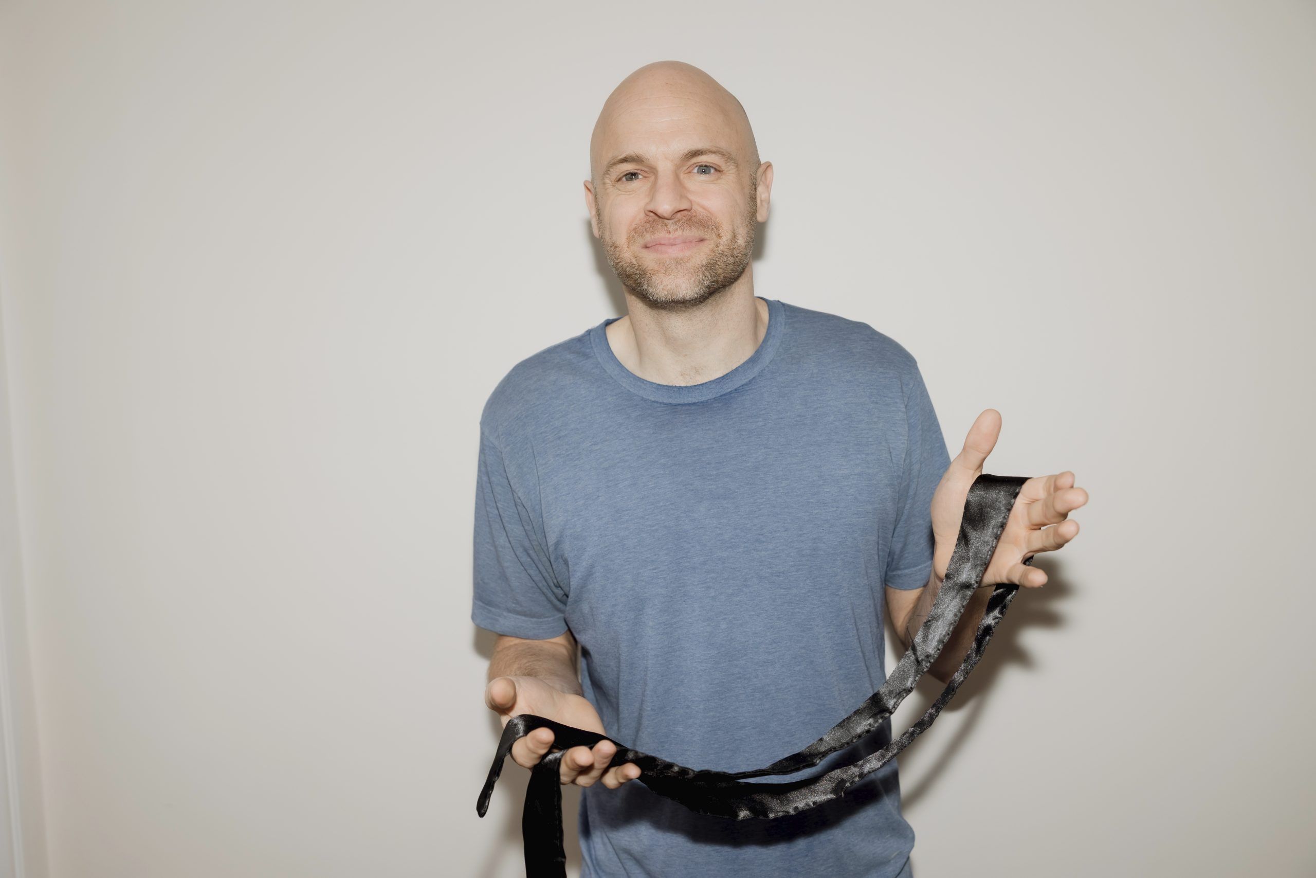 acrobat Chris Marx wearing light grey/blue t shirt holding a black blindfold against a white background