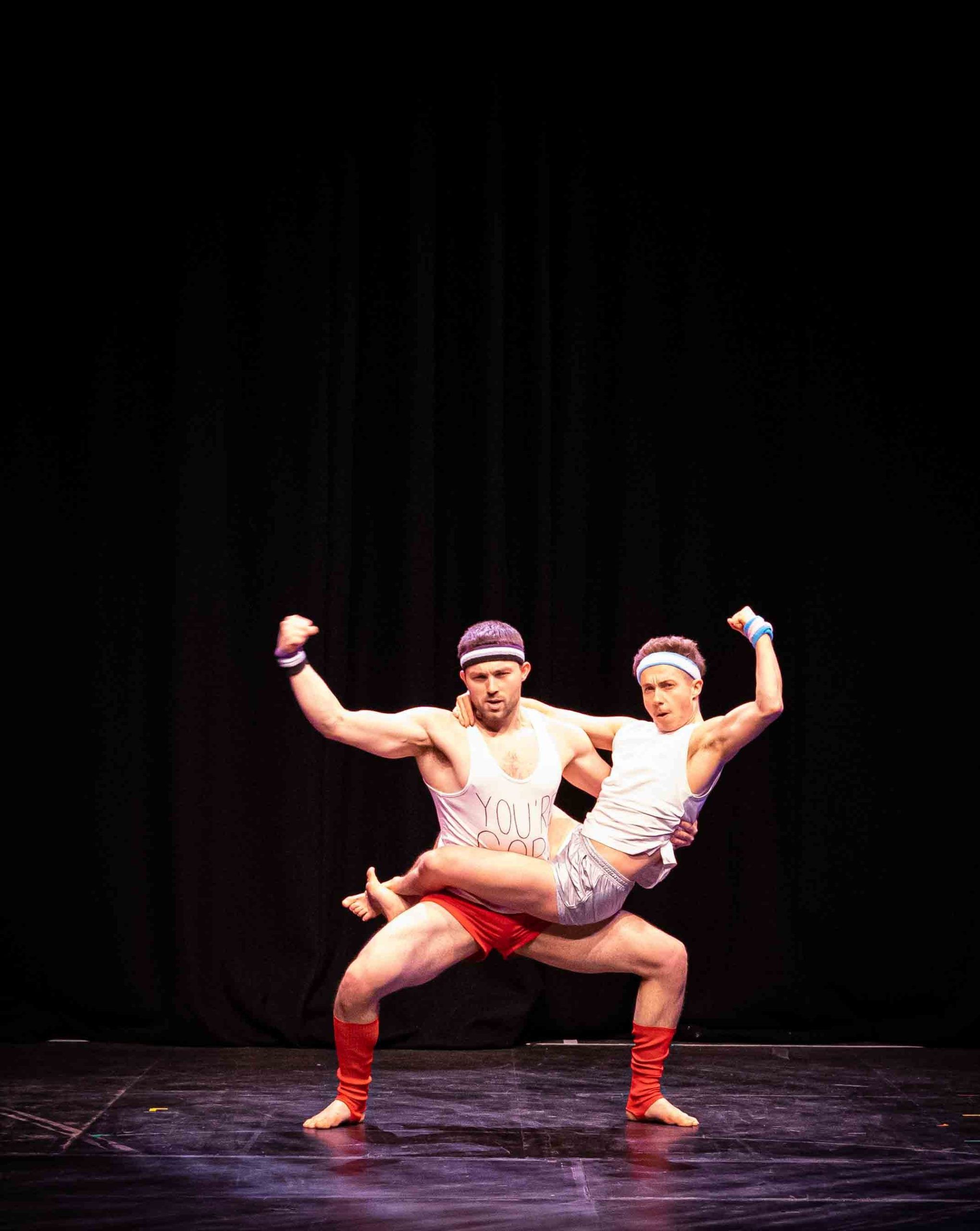 ORan Longmuir & Eric Munday performing a hands-free two-high acrobatic pose in red shorts and a white vests